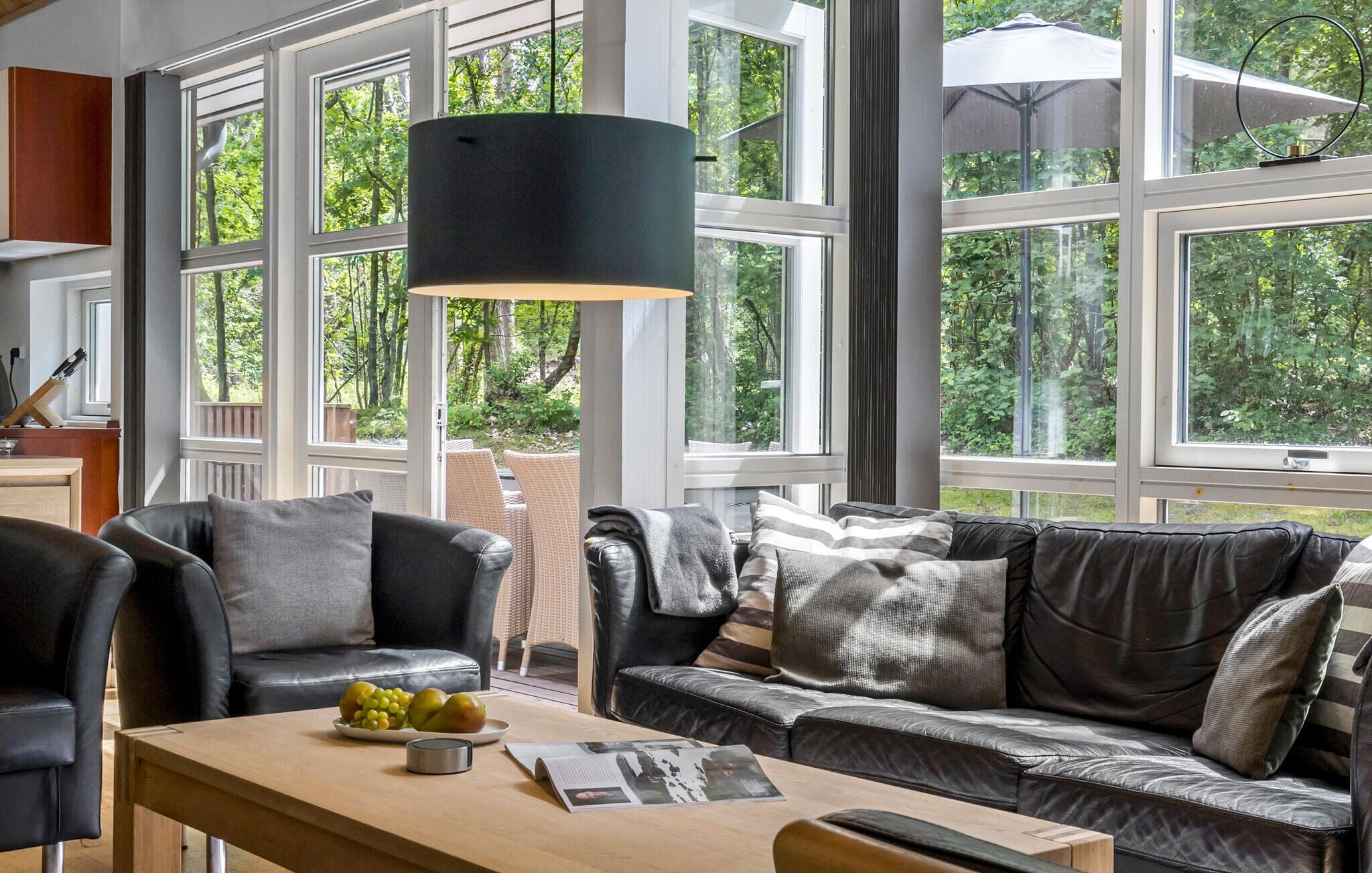 A modern living room with a leather sofa, a wooden coffee table with fruits and a magazine, and large windows offering a view of greenery.