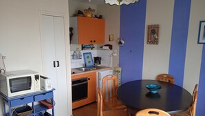 Een kleine keuken met een magnetron, broodrooster en oven. Er staat een eettafel met stoelen en een blauwe kom erop.