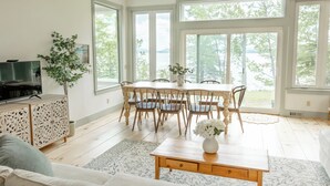 A spacious living room with a large dining table, wooden chairs, a TV on a cabinet, and large windows offering a view of the outdoors. - Designer Oceanfront, Views of Acadia National Park, Near Bar Harbor (Trenton)