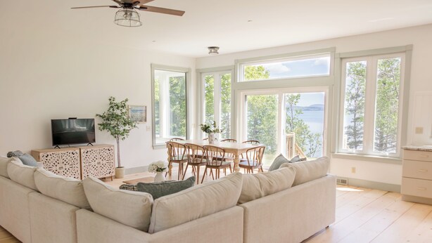 A spacious living room with a large sofa, a dining table with chairs, a television, and large windows offering a view of the outdoors. - Designer Oceanfront, Views of Acadia National Park, Near Bar Harbor (Trenton)