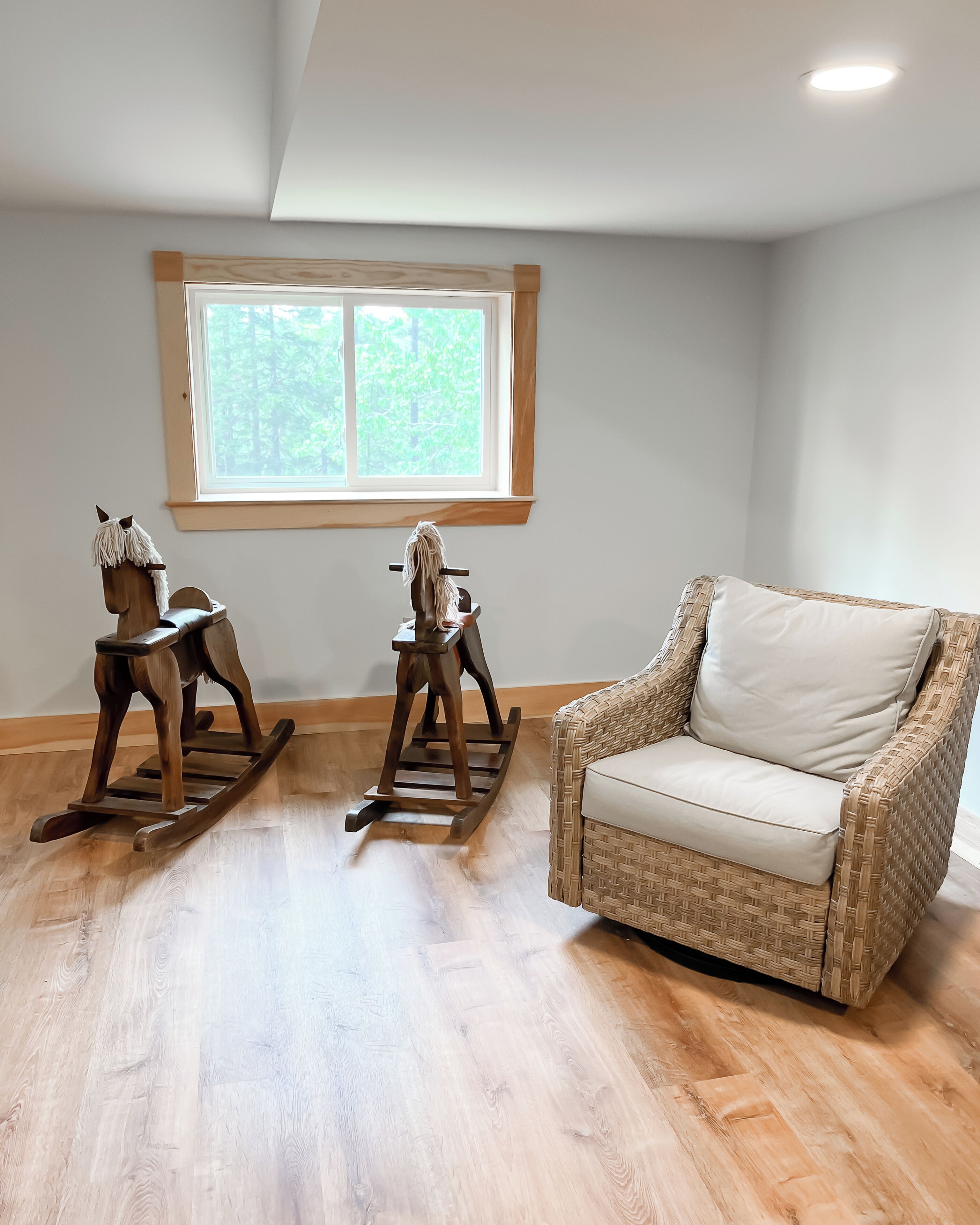 A room with a wicker chair, two wooden rocking horses, and a window with a view of trees.