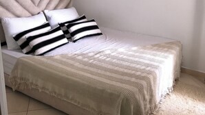 Ein ordentlich bezogenes Bett mit einem gestreiften Kopfkissen und einer strukturierten Decke, ein schlichter Kopfteil und ein gefliester Boden.