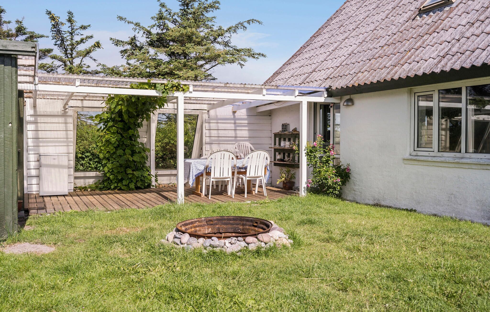 Une terrasse couverte avec une table et des chaises, un foyer extérieur et un jardin, avec une maison en arrière-plan.