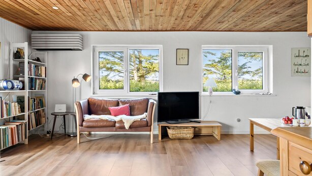 A cozy living room with a wooden ceiling, a brown leather sofa, a TV, a bookshelf, and large windows with a view of trees. - Nice home in Harboøre with WiFi (Harboøre)