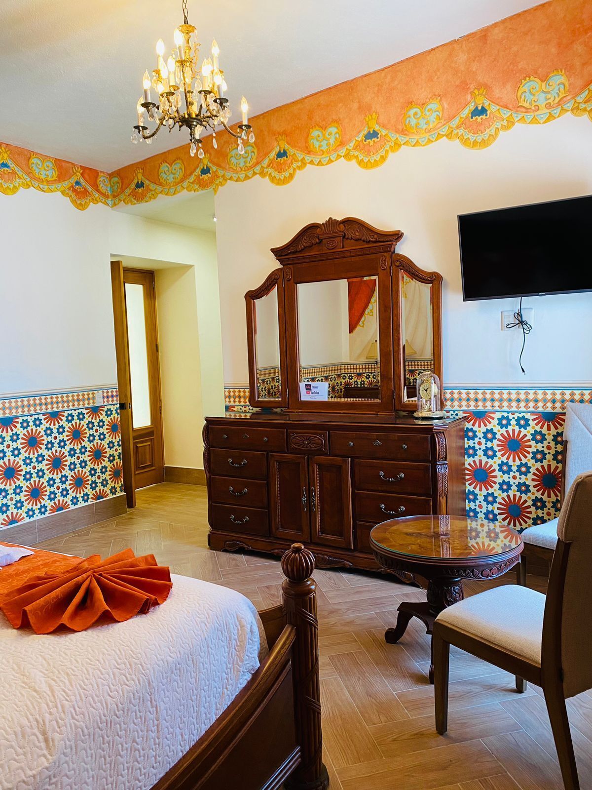 A hotel room with a bed, a wooden dresser, a round table, a chair, a chandelier, a TV mounted on the wall, and patterned walls.