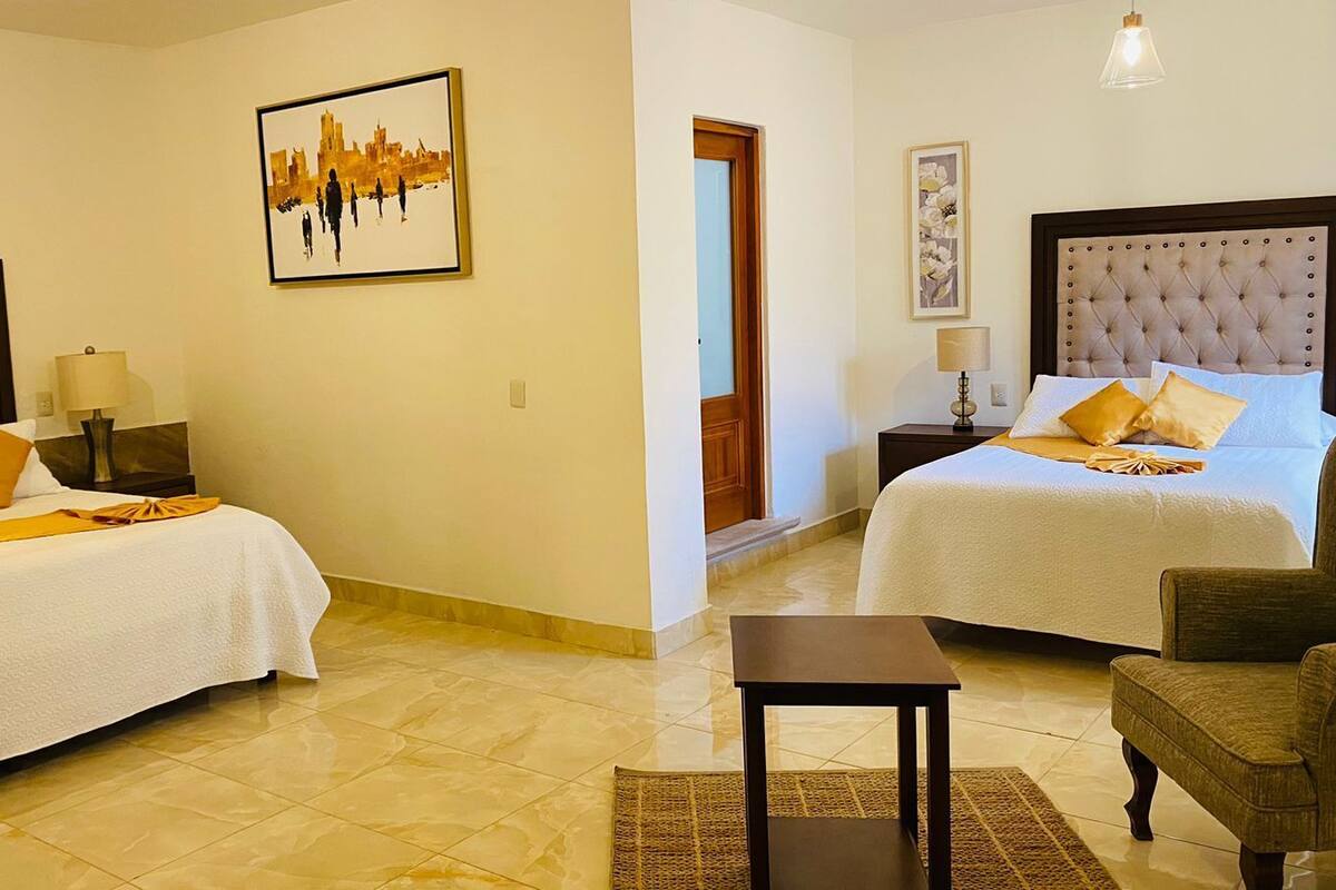 Habitación de hotel con dos camas, una mesita y una silla. Hay un cuadro en la pared y una puerta que da a otra habitación.