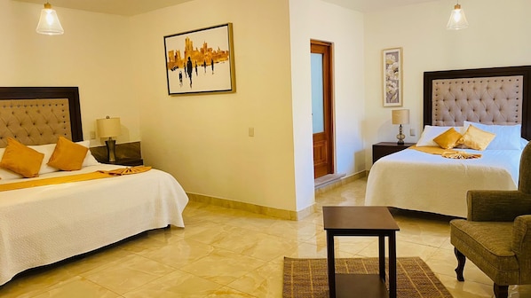 Une chambre d’hôtel avec deux lits, une petite table et une chaise. Il y a un tableau encadré au mur et une porte qui donne sur une autre pièce.
