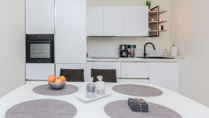 Una cucina moderna con mobili bianchi, un forno nero, una macchina da caffè e un lavello. C'è un tavolo con una ciotola di frutta, una candela e una pila di libri.