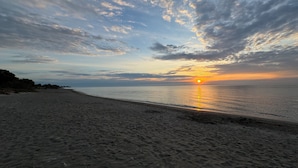 A beach with a sunset over the ocean, sandy shore, and distant trees.