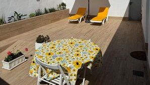En solbeskinnet terrasse med et rundt bord dækket af et blomstret dug, to gule liggestole og en stor parasol.