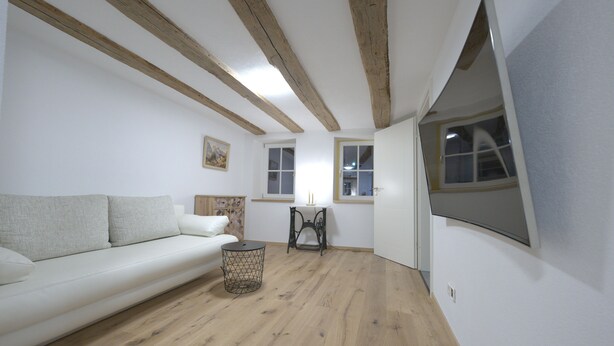 Une pièce avec un plancher en bois, des murs blancs, un canapé blanc, une petite table ronde et une fenêtre donnant sur un beau paysage.