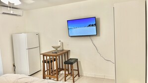 Un dormitorio con una cama, ventilador de techo, televisor en la pared, una pequeña mesa de madera con un tazón y una botella, y un refrigerador blanco.