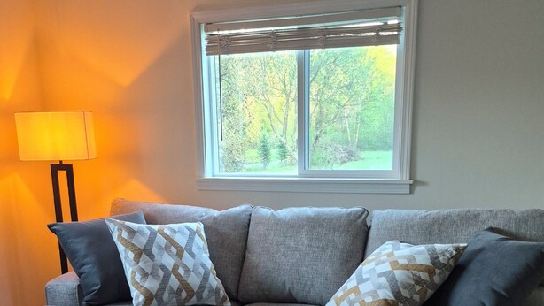 A grey sofa with patterned cushions, a patterned rug, a window with blinds, and a floor lamp. - Peaceful Vacation Home Near Wisconsin Delles with the pond on 80 Wooded Acres. (Lyndon Station)