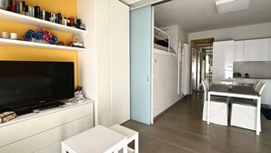 Ein modernes Wohnzimmer mit Flachbildfernseher, weißen Möbeln, einem Bücherregal und einer Küchenzeile.
