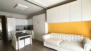 Eine moderne Küche mit weißen Schränken, einem Esstisch mit Stühlen und einem weißen Sofa an einer gelben Wand.