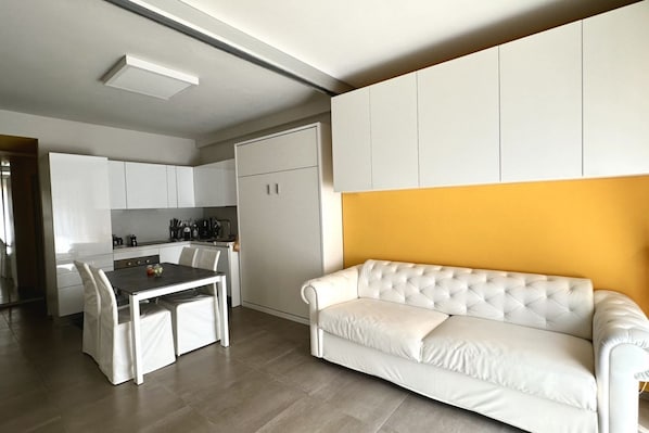 A modern kitchen with white cabinets, a dining table with chairs, and a white sofa against a yellow wall.