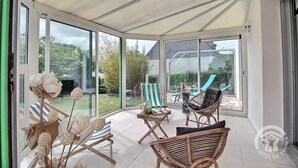 Une véranda dotée de grandes portes vitrées, de meubles en osier et offrant une vue sur un jardin ainsi que sur un espace extérieur aménagé pour s’asseoir.