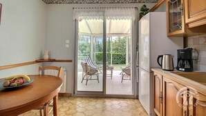 Une cuisine compacte avec un coin repas, un réfrigérateur, une machine à café et une vue sur un jardin, accessible par une porte-fenêtre coulissante.