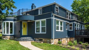 A two-story house with a stone foundation, a yellow front door, and a balcony with black railings. - Spacious Home! Game Room, Swing Set, Deck, Yard, Outdoor Fun + Sleeps 16 (Merriam)