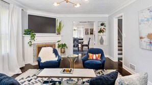 A spacious living room with a fireplace, two blue armchairs, a coffee table, and a modern chandelier. - Spacious Home! Game Room, Swing Set, Deck, Yard, Outdoor Fun + Sleeps 16 (Merriam)
