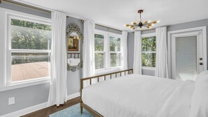A bedroom with a large bed, white curtains, a chandelier, and a view of the outdoors. - Spacious Home! Game Room, Swing Set, Deck, Yard, Outdoor Fun + Sleeps 16 (Merriam)