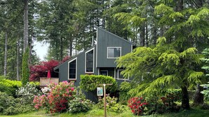 A modern two-story house with a sloped roof, surrounded by lush greenery and flowering plants. - Modern Alderbrook pet-friendly retreat.   (Union)