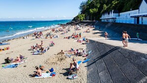 A crowded beach with people sunbathing and swimming, a lifeguard tower, and a rocky shoreline.