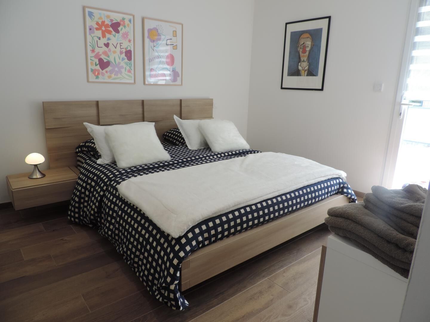 A bedroom with a wooden bed, white pillows, a patterned bedspread, a nightstand with a lamp, and artwork on the walls.