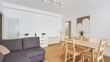 A modern living room with a grey sofa, wooden dining table with chairs, white cabinets, and a large mirror.