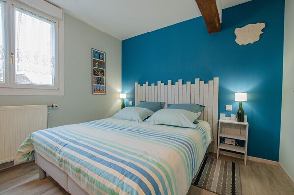 A bedroom with a bed, two bedside tables, a wall-mounted shelf, a radiator, and a window with curtains.