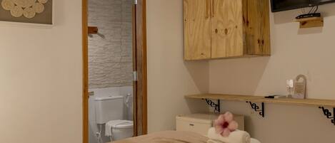 A bedroom with a wooden wardrobe, a TV mounted on the wall, a bedside table with a remote control, and a towel on the bed.