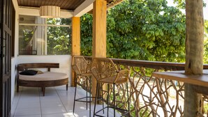 Een overdekt terras met een houten bank en rieten stoelen, omgeven door een houten leuning en weelderige begroeiing.