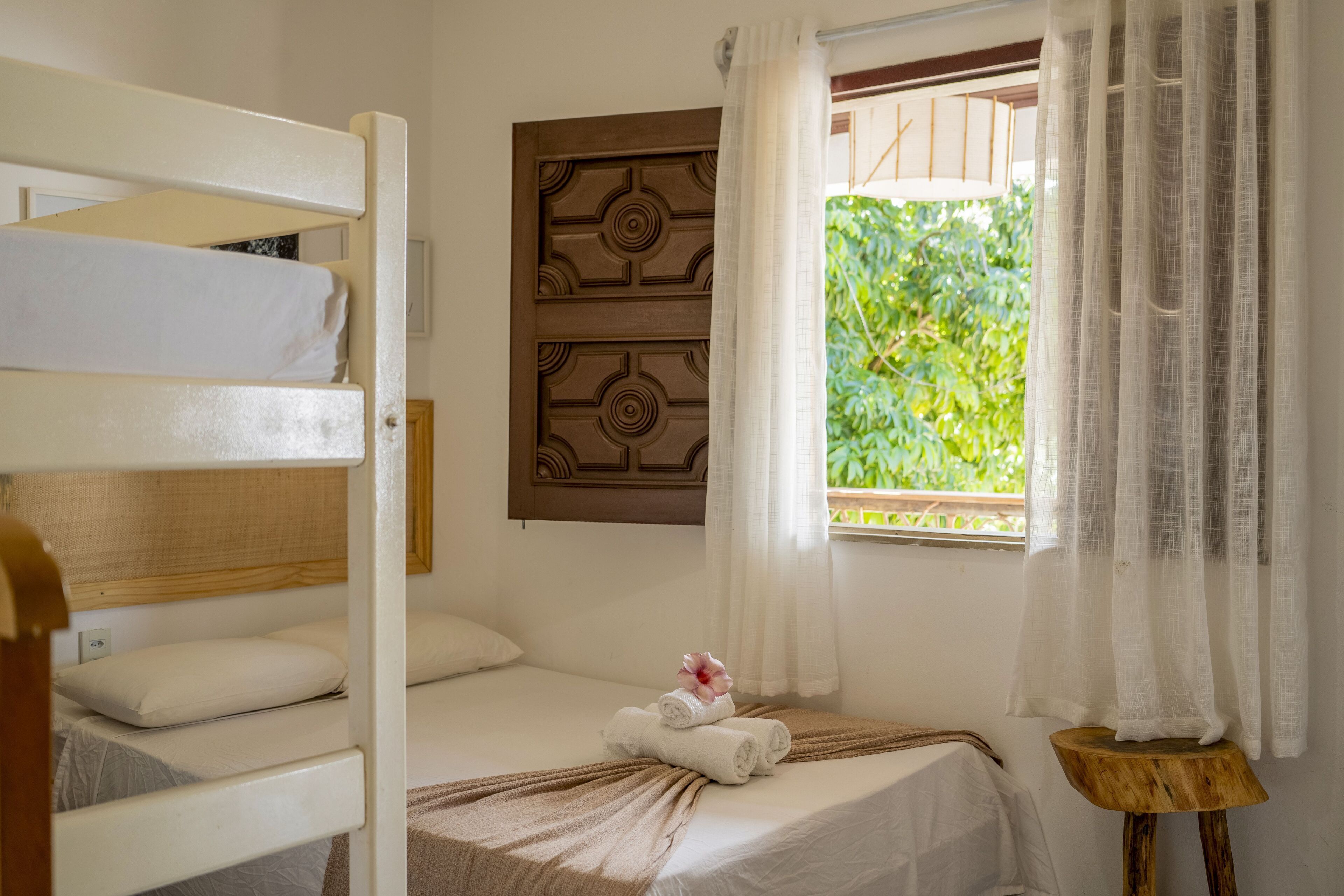 Une chambre avec un lit superposé, un lit simple avec une literie bien faite et une serviette, un petit tabouret en bois et une fenêtre avec des rideaux.