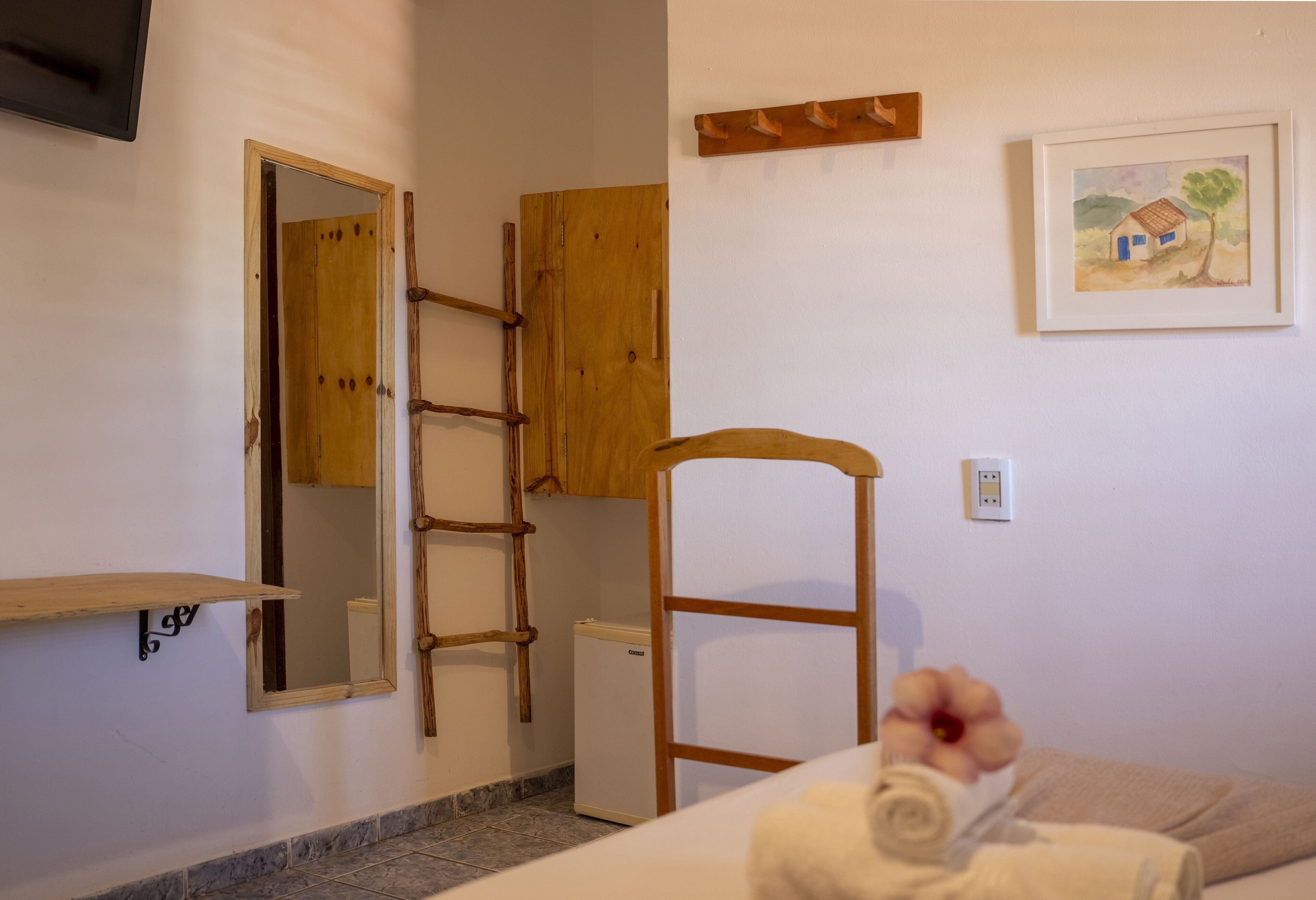 Ein Schlafzimmer mit einem Bett, einem Holzstuhl, einem kleinen Kühlschrank, einer Holzleiter, einem Regal, einem Spiegel und einem Bild an der Wand.