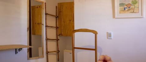 Un dormitorio con cama, una silla de madera, un refrigerador pequeño, una escalera de madera, una repisa, un espejo y un cuadro en la pared.