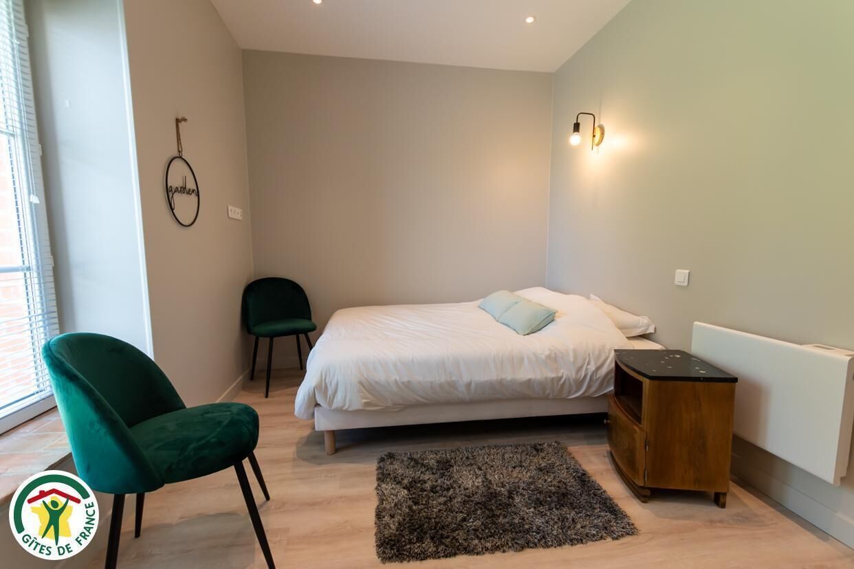 A bedroom with a bed, a green chair, a small wooden nightstand, and a wall-mounted light fixture.
