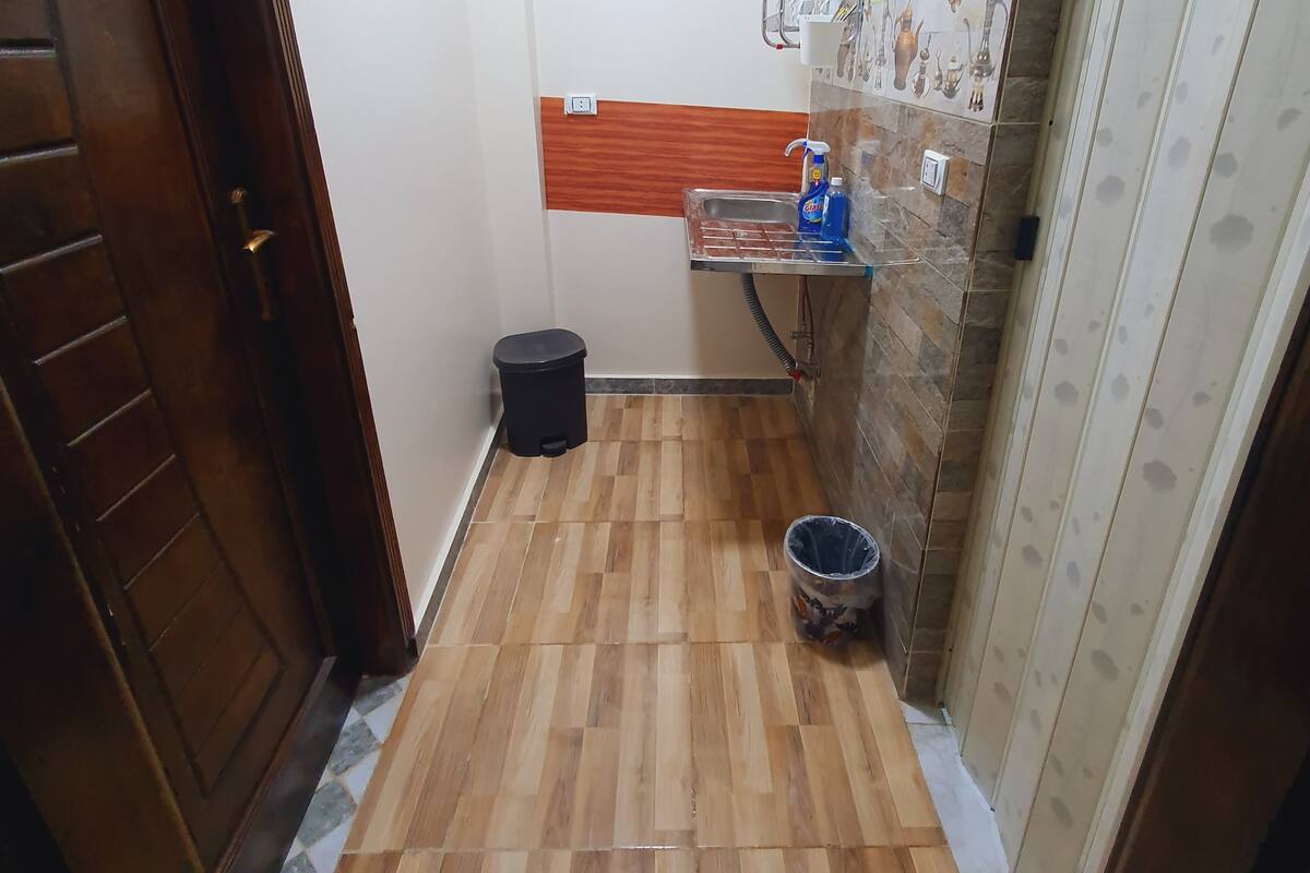 Une petite salle de bain étroite, avec un plancher en bois, un lavabo doté d’un distributeur de savon, une poubelle et un espace douche.