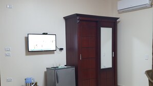 A room with a television mounted on the wall, a wardrobe, a refrigerator, a small stool, and a patterned rug. - Andy guest house  (Giza)
