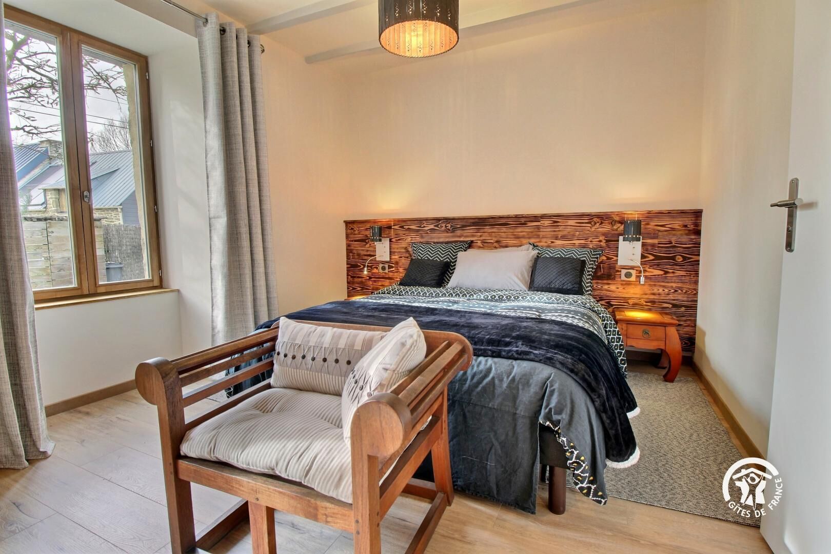 Una camera da letto con una testiera in legno, un letto con biancheria a motivi, un comodino, una poltrona con cuscini e una finestra con tende.