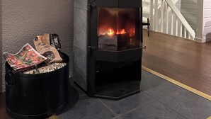 A wood-burning stove with a potted plant on top, a fire burning inside, and a bag of wood nearby.