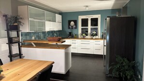 A modern kitchen with white cabinets, a wooden countertop, and a central island. There is a refrigerator on the right, a sink with a window above it, and a plant on the counter.