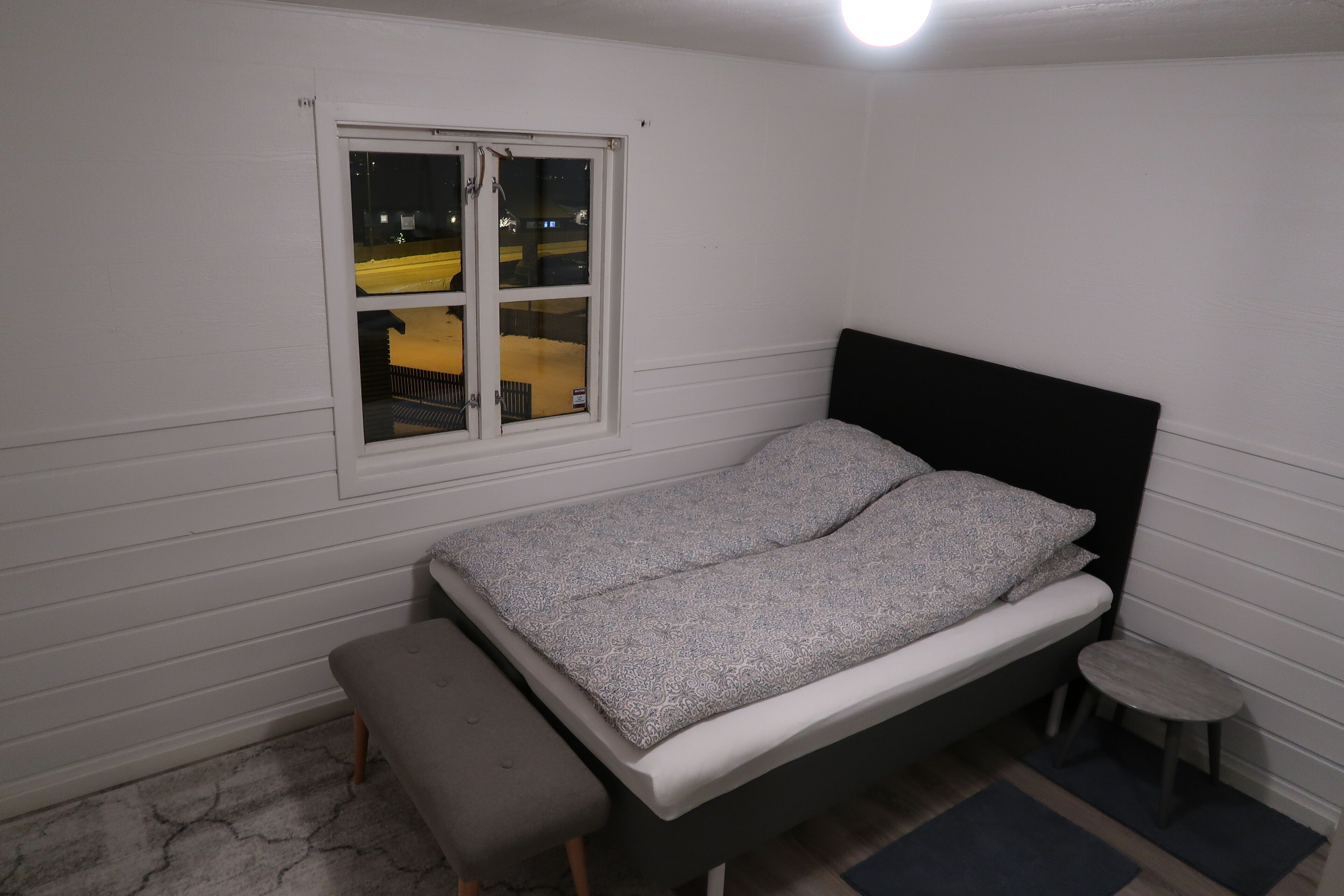 Ein Einzelbett mit Kopfteil, eine Bank, ein kleiner Tisch und ein Fenster mit Blick auf ein gelbes Gebäude bei Nacht.
