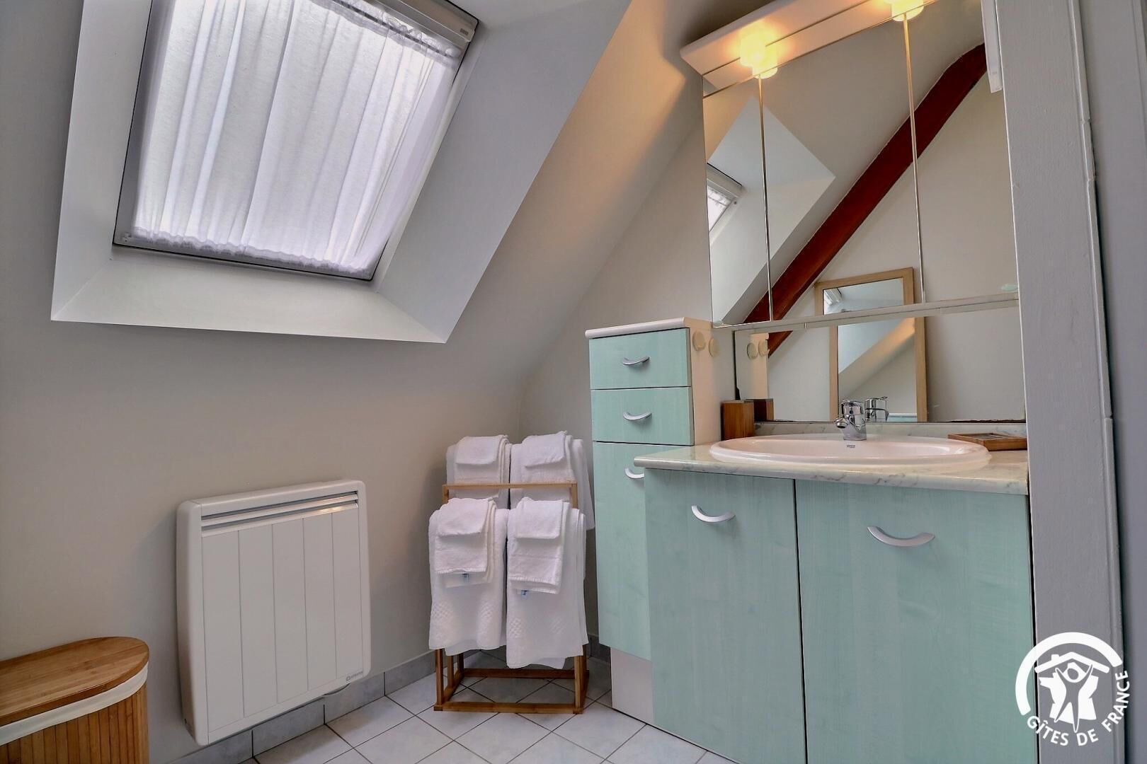 Ein modernes Badezimmer mit Dachfenster, einem weißen Heizkörper, einem Waschbecken mit Spiegel und einem hölzernen Hocker mit Handtüchern.