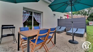 Un patio al aire libre con una mesa de madera y sillas azules, un gran paraguas verde y una zona de sofás con cojines.