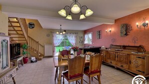 Un salón amplio con escalera de madera, comedor con mesa y sillas, cocina con mueble de madera y un gran ventanal con cortinas.