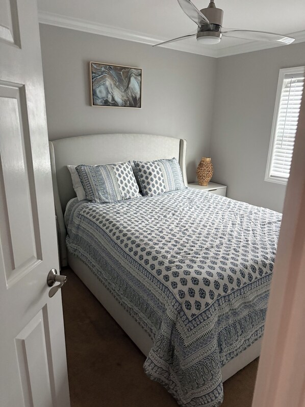 Un dormitorio con cama, ventilador de techo, un cuadro en la pared y una ventana con persianas.