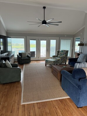 A spacious living room with a ceiling fan, large windows, and a flat-screen TV.
