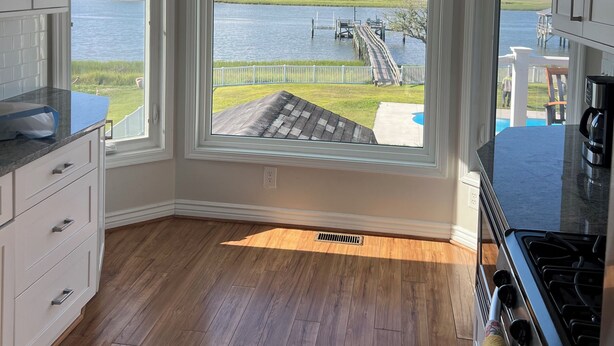 A kitchen with large windows overlooking a lake, wooden flooring, and white cabinetry. - Waterway Home with Private Dock, Private Pool and Fenced in yard (Ocean Isle Beach)