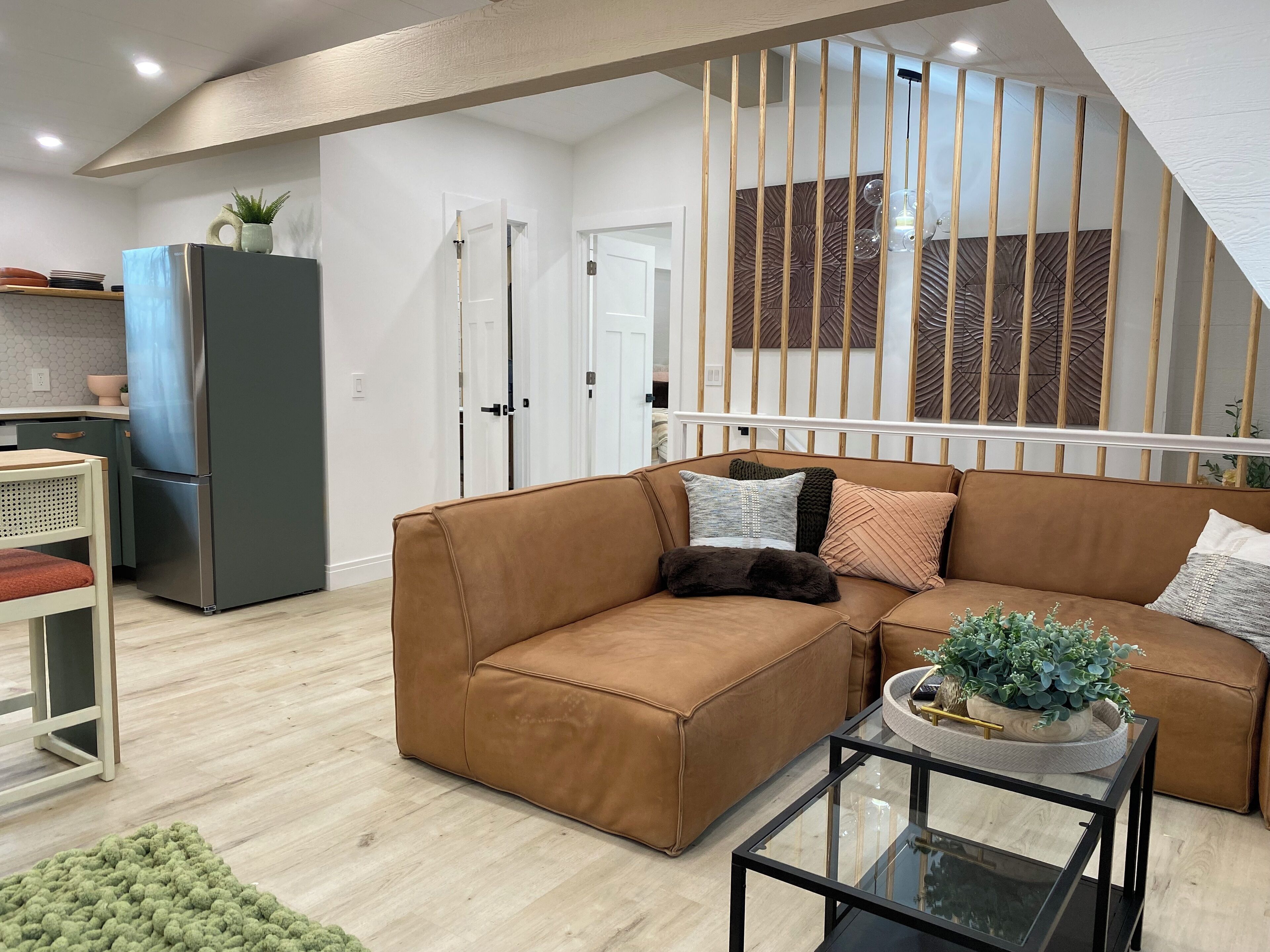 A modern living room with a brown leather sectional sofa, a glass coffee table, and a kitchen area with a refrigerator and cabinets.