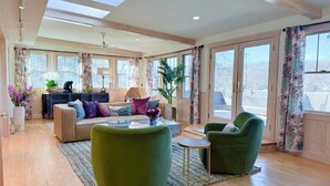 A spacious living area with a sofa, armchairs, a coffee table, and a dining table. There are large windows with curtains, a ceiling fan, and a skylight.
